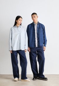 Lichtblauwe oversized button-up shirt, donkere spijkerjas, geruite shirt eronder, marineblauwe wijde broek, witte sneakers en zwarte instappers.