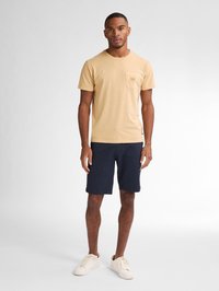 Tricou cu mânecă scurtă, de culoare bej deschis, cu buzunar în față, asortat cu pantaloni scurți de culoare albastru închis și adidași albi. Textură din bumbac fin.