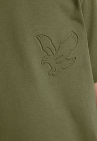 Oliven grøn bomulds t-shirt med et præget ørnerlogo på venstre side. Stoffet har en glat tekstur og en let tætsiddende pasform.
