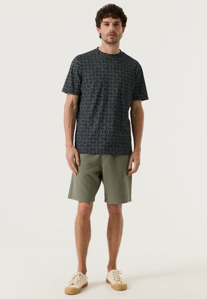 Man die staat in een donkergrijs T-shirt met patroon, olijfgroene korte broek tot aan de knie en gebroken witte sneakers tegen een effen lichte achtergrond.