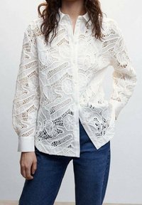 White lace shirt with detailed cut-out patterns, long sleeves, and a buttoned front. Paired with dark blue jeans.