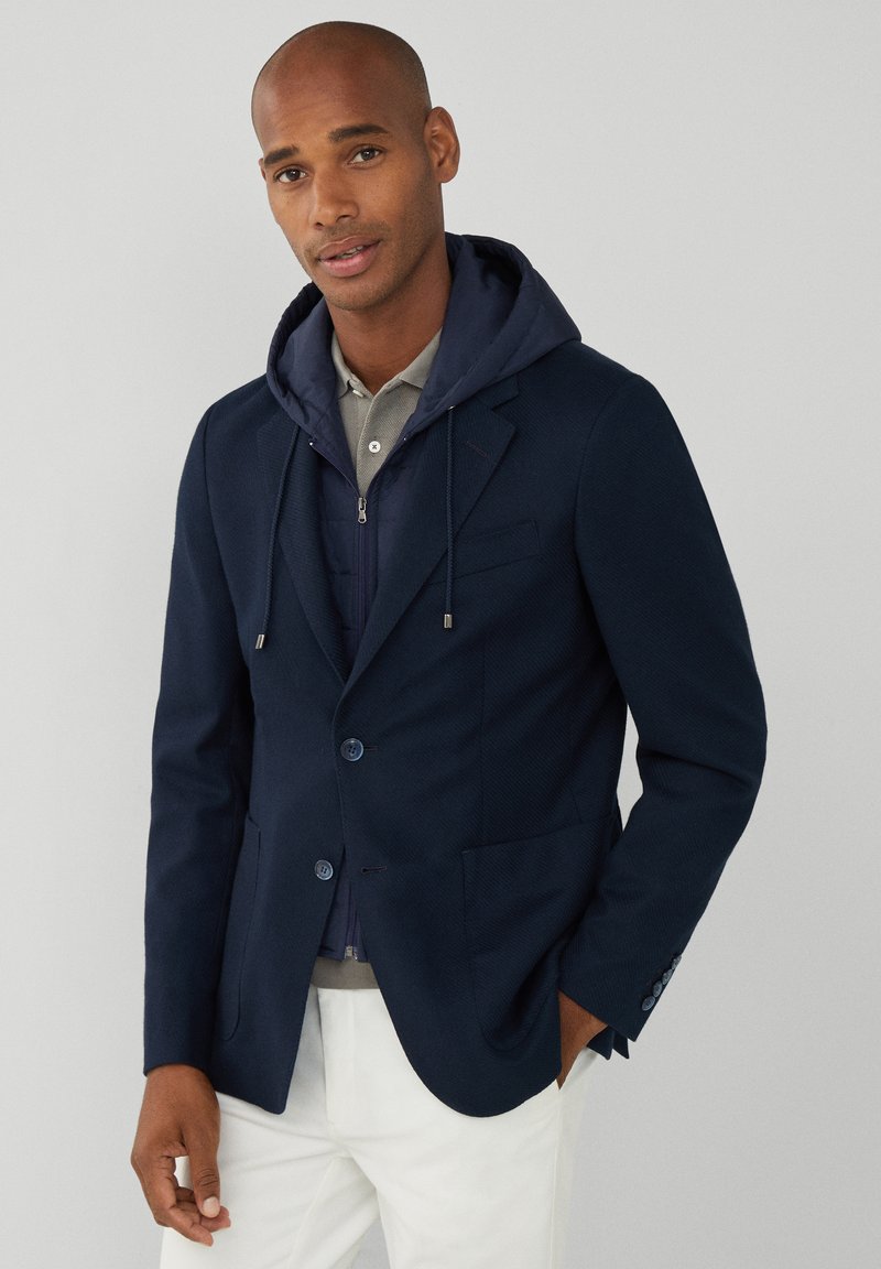 Blazer azul marino con tela texturizada, dos botones y bolsillos parche; llevado sobre una sudadera oscura con capucha y un polo gris claro; combinado con pantalones blancos.