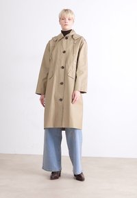 Μπεζ αδιάβροχο με κουκούλα, κουμπιά μπροστά και δύο μεγάλες πλευρικές τσέπες. Φοριέται πάνω από ένα μαύρο πουλόβερ με ψηλό λαιμό και συνδυάζεται με ελαφριά τζιν παντελόνια φαρδιάς γραμμής.