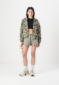Camo print bomber jacket in green tones, black crop top, light grey shorts with pockets, white socks, and beige sneakers. Minor accessories included.
