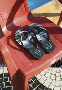 Tongs à motif noir et gris avec des brides noires placées sur une chaise en plastique rouge sur un sol en carrelage mosaïque.