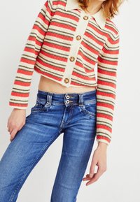 Cardigan corto a righe in corallo, crema e oliva con colletto beige e grandi bottoni, abbinato a jeans blu aderenti.
