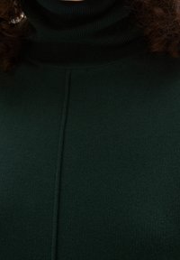 Kudrnaté vlasy viditelné nad tmavě zeleným ribbovým rolákem s centrálním svislým švem na látce.