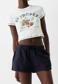 Bílá zkrácená tričko s barevnými ovocnými grafikami a textem, v kombinaci s tmavě modrými šortkami na šňůrku vyrobenými z lehkého materiálu.