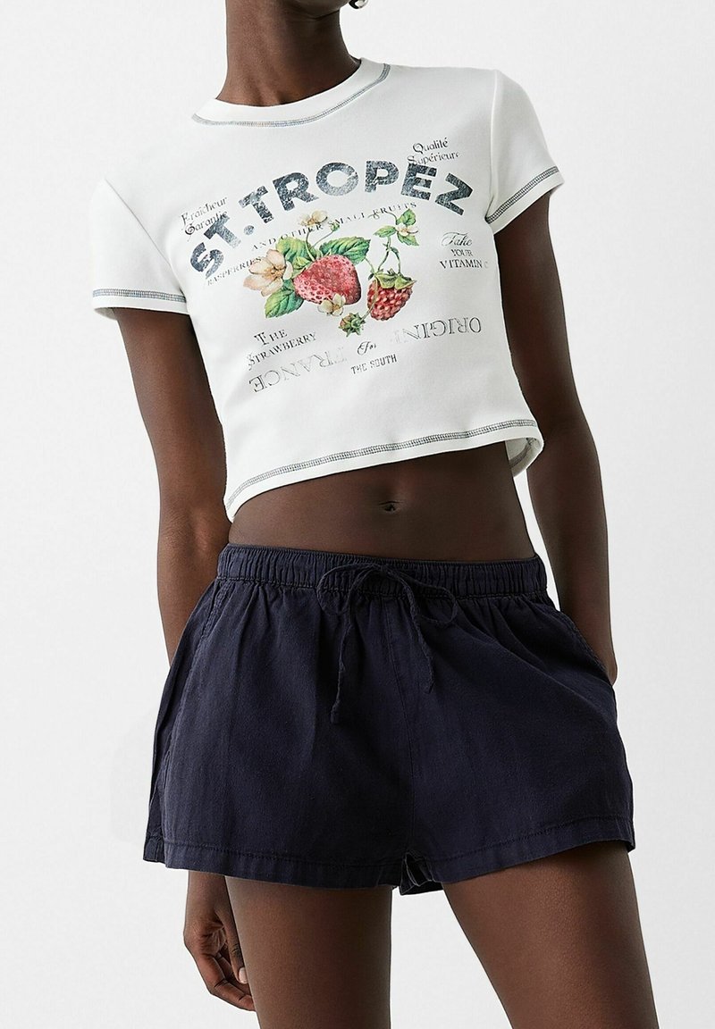 Bílá zkrácená tričko s barevnými ovocnými grafikami a textem, v kombinaci s tmavě modrými šortkami na šňůrku vyrobenými z lehkého materiálu.