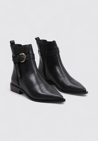 Black leather ankle boots with pointed toes, flat soles, and a side strap with a gold circular buckle. Smooth texture, minimalistic design.