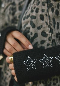 Černý pletený manžetový návlek se třemi stříbrnými hvězdičkami, v kombinaci se šedou bundou s leopardím potiskem. Na ruce zdobený zlatým prstenem.