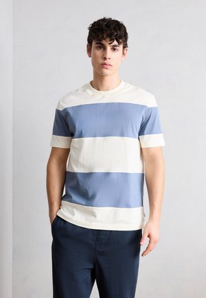 Mladý muž stojí čelom vpred, oblečený v tričku s krátkym rukávom s širokými modro-bielymi vodorovnými pruhmi a tmavými nohavicami.