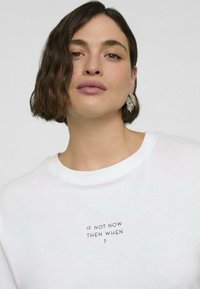 Weißer Baumwollpullover mit kurzen Ärmeln, mit schwarzem Text "WENN NICHT JETZT, WANN DANN?" auf der Vorderseite bedruckt. Glatte Textur, Rundhalsausschnitt.