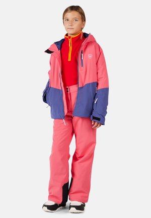 Enfant portant une veste de ski rose et violette, un haut zippé rouge et un pantalon de neige rose avec des bottes noires et blanches, se tenant devant un fond blanc.