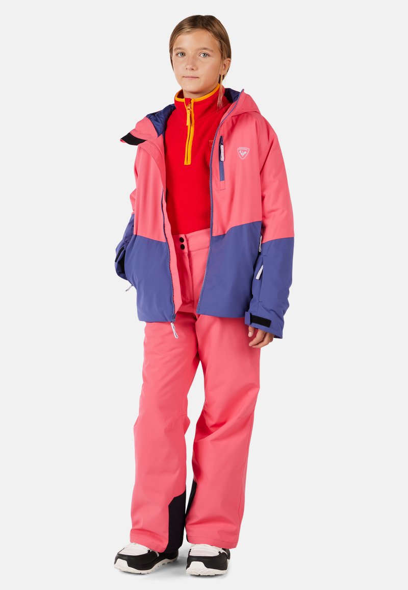 Dítě oblečené v růžové a fialové lyžařské bundě, červeném zipovém svetru a růžových sněhových kalhotách s černobílými botami, stojí na bílém pozadí.