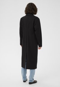 Personne aux cheveux bouclés portant un long manteau noir, un jean bleu clair et des chaussures noires, debout de dos face à la caméra.