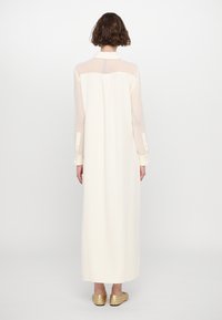 Largo vestido de crema con una sección superior transparente, un cuello y puños con botones. Presenta un ajuste relajado con una textura suave y un diseño minimalista.