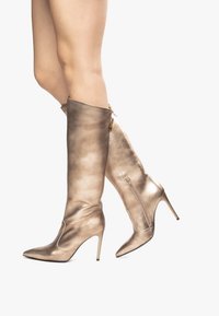 Bottes dorées métalliques jusqu'au genou avec un bout pointu et un talon aiguille. Présentent un design élégant et une fermeture à glissière sur le côté intérieur.