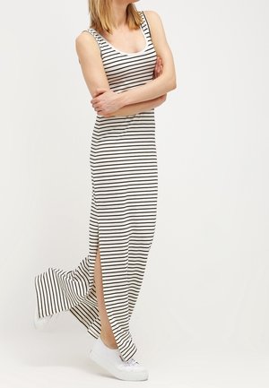 Kobieta stojąca z założonymi rękami, ubrana w białą sukienkę maxi bez rękawów w czarne paski oraz białe trampki, na białym tle.