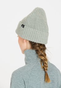 Grå stickad mössa med hög krona och uppvikt brätte, med en liten svart logotypetikett. Bärs tillsammans med en grå topp med hög krage.