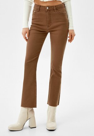 Vrouw draagt bruine rechte broek met zijslitsen en witte enkellaarsjes met hak, staand tegen een effen achtergrond.