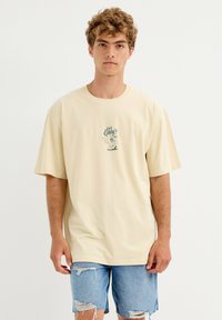 Joven con cabello rizado, vistiendo una camiseta beige de gran tamaño con un diseño gráfico y pantalones cortos de mezclilla con las rodillas rasgadas, de pie frente a un fondo liso.