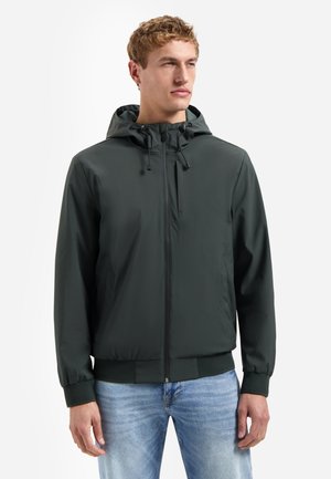 Junger Mann trägt eine dunkelgrüne Kapuzenjacke mit Reißverschluss und hellblaue Jeans, steht vor weißem Hintergrund und schaut nach rechts.