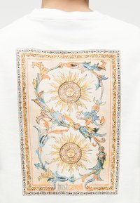 Λευκό πίσω μέρος πουκάμισου με περίτεχνο ορθογώνιο σχέδιο που απεικονίζει δύο κίτρινες ηλιακές μορφές, περιτριγυρισμένες από μπλε και πορτοκαλί διακοσμητικά λουλουδένια μοτίβα.