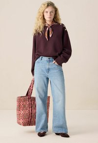 Junge Frau mit langen blonden Haaren, die einen lila Pullover, hellblaue weite Jeans, ein gemustertes Halstuch trägt und eine große Blumentasche hält.