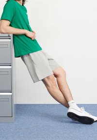 Zelené bavlněné tričko, šedé šortky, bílé tenisky, postaveno proti skříni na dokumenty s pozadím světle modrého koberce.