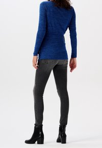 Kobieta ubrana w dopasowaną niebieską bluzkę z długim rękawem, szare jeansy skinny i czarne botki na obcasie, stojąca na tle białym.