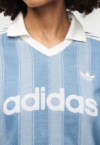 Μπλε φανέλα Adidas με κάθετες ρίγες, λευκό γιακά και λευκό λογότυπο. Το υλικό φαίνεται μαλακό με ομαλή υφή.