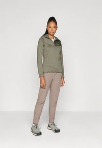 Casaco verde-oliva com fecho de correr e capuz, dois bolsos frontais e detalhes em laranja, combinado com calças castanho-claro e sapatilhas de caminhada cinzentas.