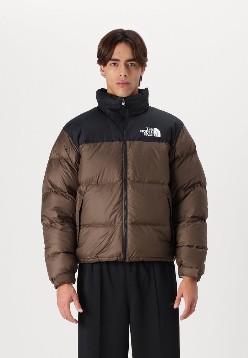 Jachetă puf maro și neagră cu guler înalt, dotată cu un fermoar frontal și logo-ul North Face pe piept. Materialul are o textura lucioasă.