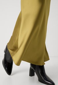 Black heeled boots with a pointed toe paired with a smooth, olive-green satin skirt, featuring an A-line silhouette and slight drape.