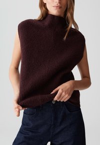 Bordeaux mouwloze trui van ribgebreide stof, met een hoge kraag en een losse pasvorm, gecombineerd met donkerblauwe jeans.