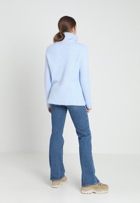 Kvinna som står vänd bort, klädd i en ljusblå polotröja, blå jeans och beige klumpiga sneakers mot en enfärgad bakgrund.