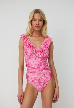 Kobieta w różowym kwiecistym jednoczęściowym kostiumie kąpielowym z falbankowym dekoltem w kształcie litery V i marszczonym paskiem, stojąca na szarym jednolitym tle.