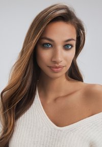 Gebreide, off-shoulder trui in zacht wit, met een textuurpatroon; lange, golvende bruine haren; opvallende blauwe ogen; zachte, natuurlijke make-up.