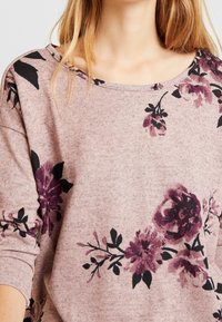 Sweatshirt rose clair avec imprimé floral dans des nuances de bordeaux et de noir, doté d'un large col et de manches longues. Texture de tissu douce.