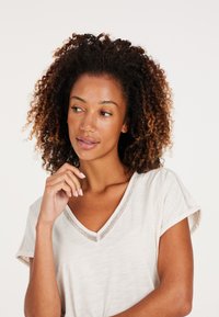 Svetlo béžový top s V-výstrihom a krátkymi rolovanými rukávmi, ktorý má textúrovaný sieťový detail okolo výstrihu. Hladká, ľahká tkanina.