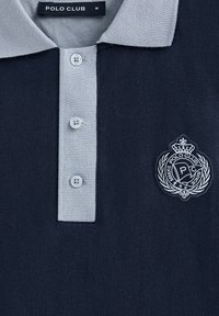 Marinblå polo skjorta med ljusgrå krage, tre-knappslå och broderad logo med en krona och kransdetalj. Storlek M.