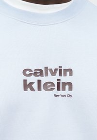 Világoskék pulóver texturált anyagból, közepén vastag sötétszürke „calvin klein” logóval és „New York City” felirattal.