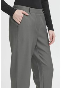 Pantalons gris sur mesure en tissu lisse, dotés d'une taille élastique et de poches latérales, présentés à la taille sur un modèle.