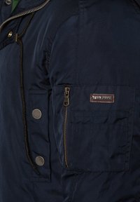 Granatowa kurtka z zamykana kieszonką i guzikami na rękawie. Wykonana z gładkiego materiału o delikatnym połysku.