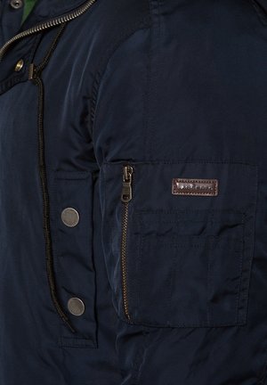Granatowa kurtka z zamykana kieszonką i guzikami na rękawie. Wykonana z gładkiego materiału o delikatnym połysku.