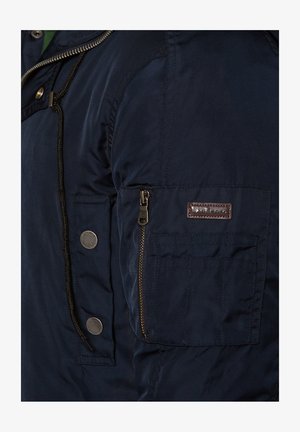 Granatowa kurtka z zamykana kieszonką i guzikami na rękawie. Wykonana z gładkiego materiału o delikatnym połysku.