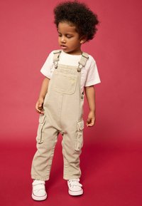 Salopette beige claire avec des bretelles réglables, poche avant et rabats latéraux, associée à une chemise blanche et des baskets blanches à velcro.