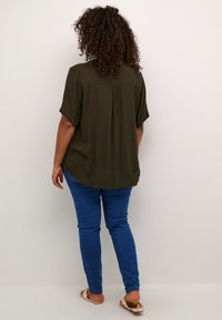 Blusa verde oliva a maniche corte con orlo arrotondato e piega sul retro; abbinata a jeans skinny blu e sandali beige.