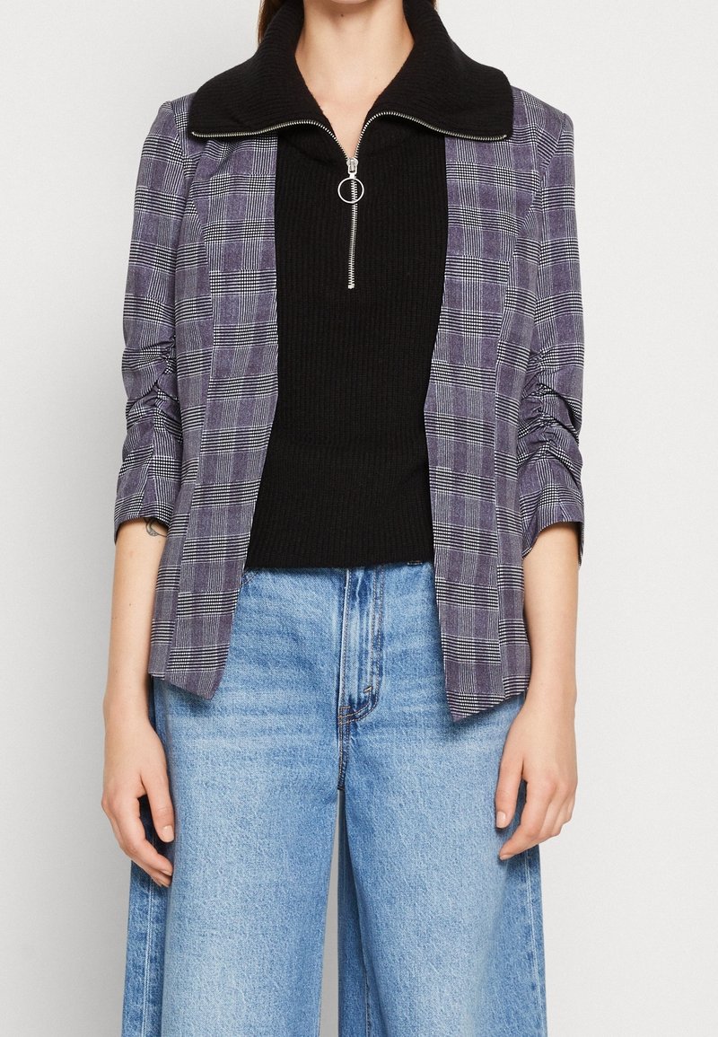 Blazer à carreaux dans des tons de violet et de gris, avec un col zippé noir. Porté sur un haut noir côtelé avec une fermeture éclair et associé à un jean à jambes larges.